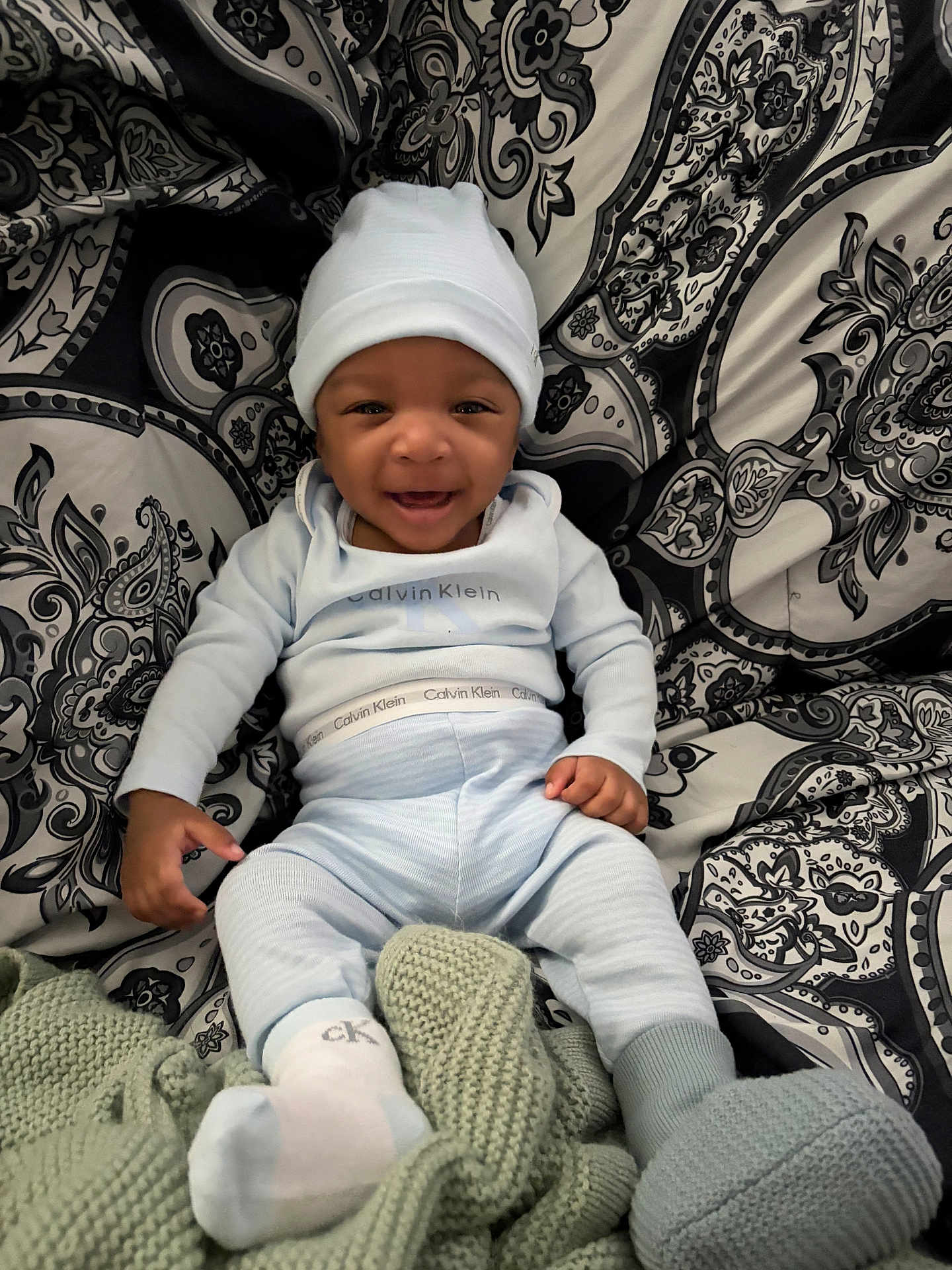 Jacai is registered to the contest to win money with this photo: baby, smiling, infant, hat, clothing, calvin_klein, pajamas, blanket, knitted, patterned_background, paisley, sitting, happy, cozy, indoor, cute, portrait, child, warm, soft