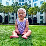 Evelyn is registered to the contest to win money with this photo: baby, child, grass, outdoor, building, palm_tree, headband, romper, barefoot, greenery, sunlight, portrait, sitting, cute, infant, summer, nature, smiling, young, daytime