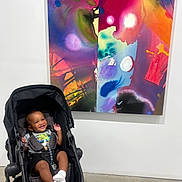 Landon joined the competition — help win amazing prizes! abstract_painting, art_gallery, baby, black_stroller, bright, casual_clothing, child, colorful, concrete_floor, happy, indoor, infant, modern, playful, portrait, seated, smiling, sock, stroller, white_wall