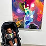baby, stroller, smiling, indoor, abstract_painting, art_gallery, concrete_floor, colorful, child, happy, portrait, seated, white_wall, modern, casual_clothing, sock, black_stroller, infant, playful, bright
