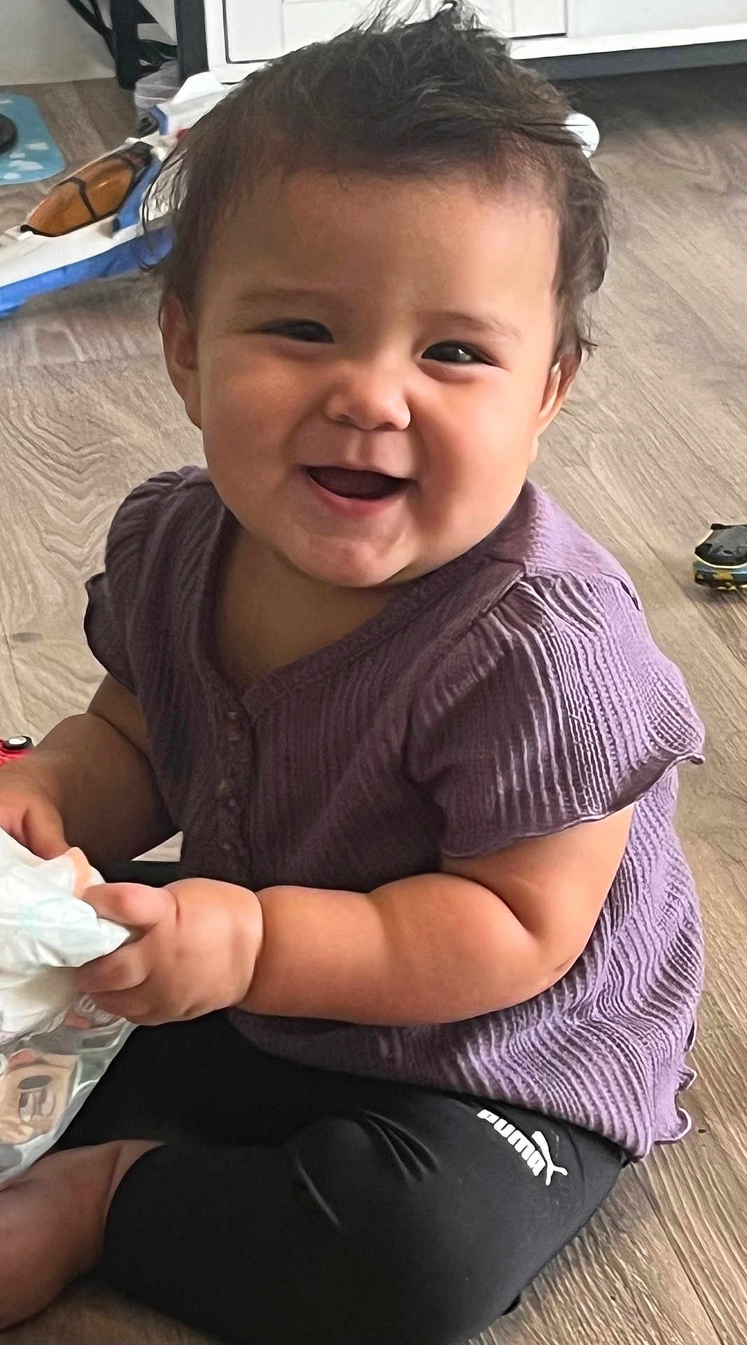 Violet is registered to the contest to win money with this photo: baby, child, smiling, purple_shirt, black_pants, puma_logo, sitting, floor, wooden_floor, toy, diaper, happy, indoor, cute, chubby_arms, short_hair, playtime, person, infant, casual_clothing