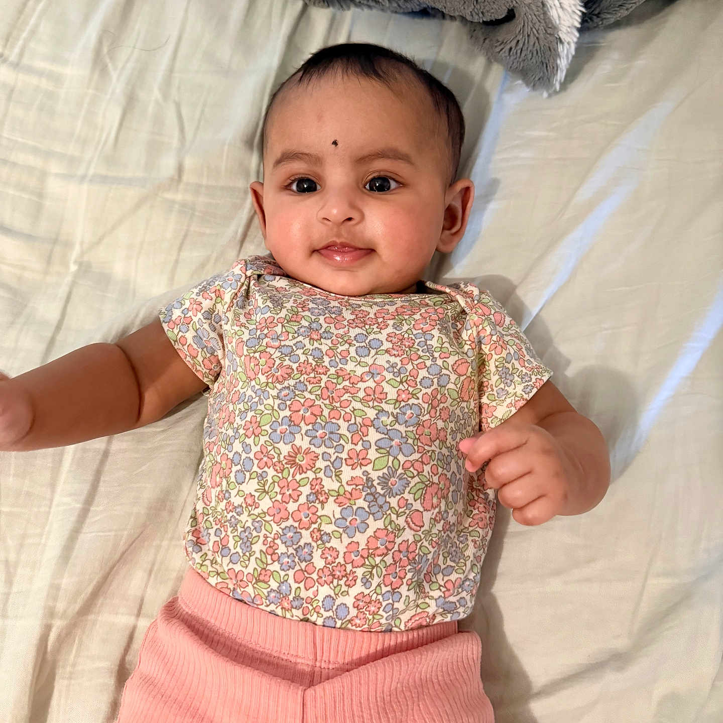 Kanika joined the competition — help win amazing prizes! baby, bed, blanket, child, cute, eyes, floral_shirt, hand, happy, head, indoors, infant, lying_down, person, pink_pants, portrait, skin, smiling, soft_texture, young_child