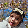blossom, blue_sky, bow, child, closeup, cute, face, flowers, headband, jacket, nature, outdoor, person, portrait, smiling, spring, toddler, tongue, tree_branch, warm_clothing