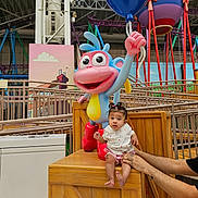 Sienna is registered to the contest to win money with this photo: baby, child, indoor, amusement_park, toy, statue, balloon, colorful, wooden_crate, adult_hand, playful, cartoon_character, person, face, seated, pink_clothing, white_shirt, hair_tied, supporting_hands, background_structure