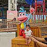 baby, child, indoor, amusement_park, toy, statue, balloon, colorful, wooden_crate, adult_hand, playful, cartoon_character, person, face, seated, pink_clothing, white_shirt, hair_tied, supporting_hands, background_structure