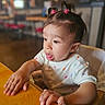 Sienna joined the competition — help win amazing prizes! toddler, child, face, hands, table, hair, ponytails, pink_clips, shirt, heart_pattern, indoor, wooden_table, curious, expression, seated, person, cafe, blurred_background, cute, exploration