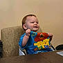 toddler, child, high_chair, blue_shirt, cartoon_characters, table, wooden_table, indoor, smiling, happy, person, baby, furniture, seated, cute, young_child, cheerful, casual_clothing, face, raising_hands