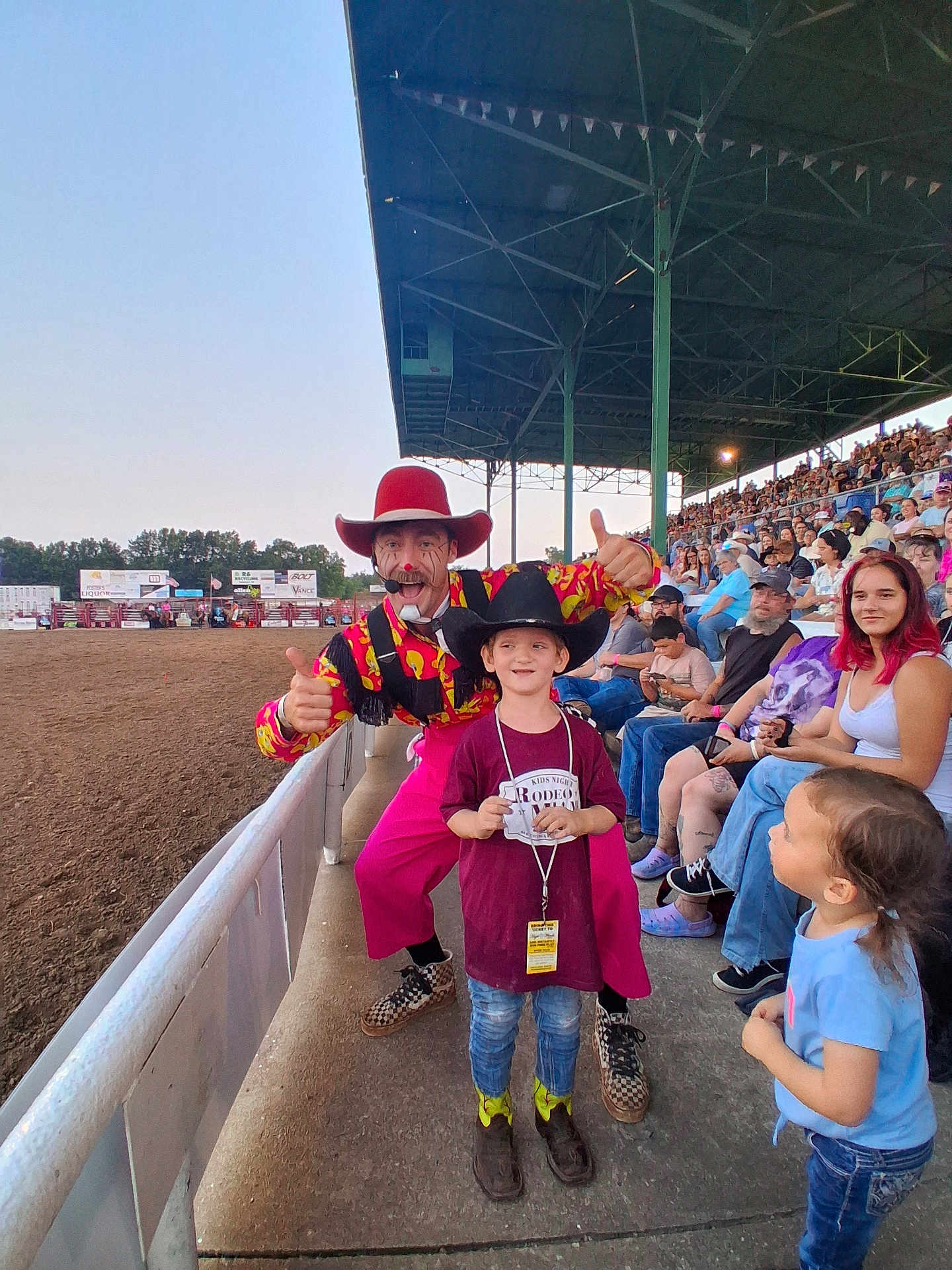 Kylo is registered to the contest to win money with this photo: child, boy, girl, rodeo_clown, cowboy_hat, crowd, stadium_seats, outdoor, arena, fence, concrete, casual_clothing, smile, thumbs_up, event, spectators, daylight, jeans, boots, hat