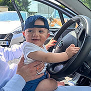 Siwaÿn participe au concours pour gagner de l'argent avec cette photo : baby, child, cap, car, steering_wheel, person, hand, seat, open_door, daylight, outdoor, smile, blue_clothing, white_shirt, ring, trees, vehicle, happy, human, lap
