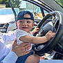 baby, child, cap, car, steering_wheel, person, hand, seat, open_door, daylight, outdoor, smile, blue_clothing, white_shirt, ring, trees, vehicle, happy, human, lap