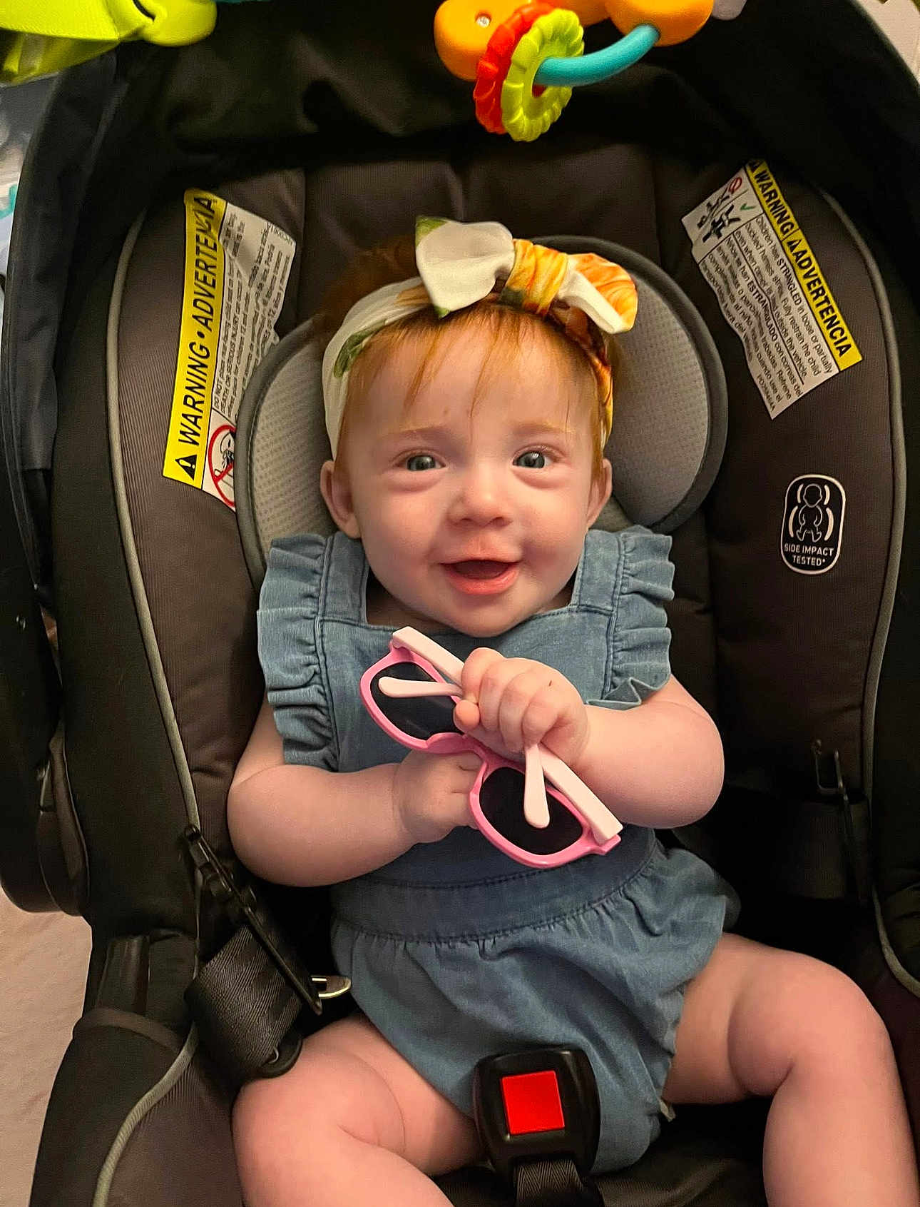 Willow is registered to the contest to win money with this photo: baby, child, car_seat, headband, pink_sunglasses, smile, happy, infant, blue_outfit, seatbelt, indoors, cute, young_child, person, holding, accessory, playful, closeup, adorable, skin