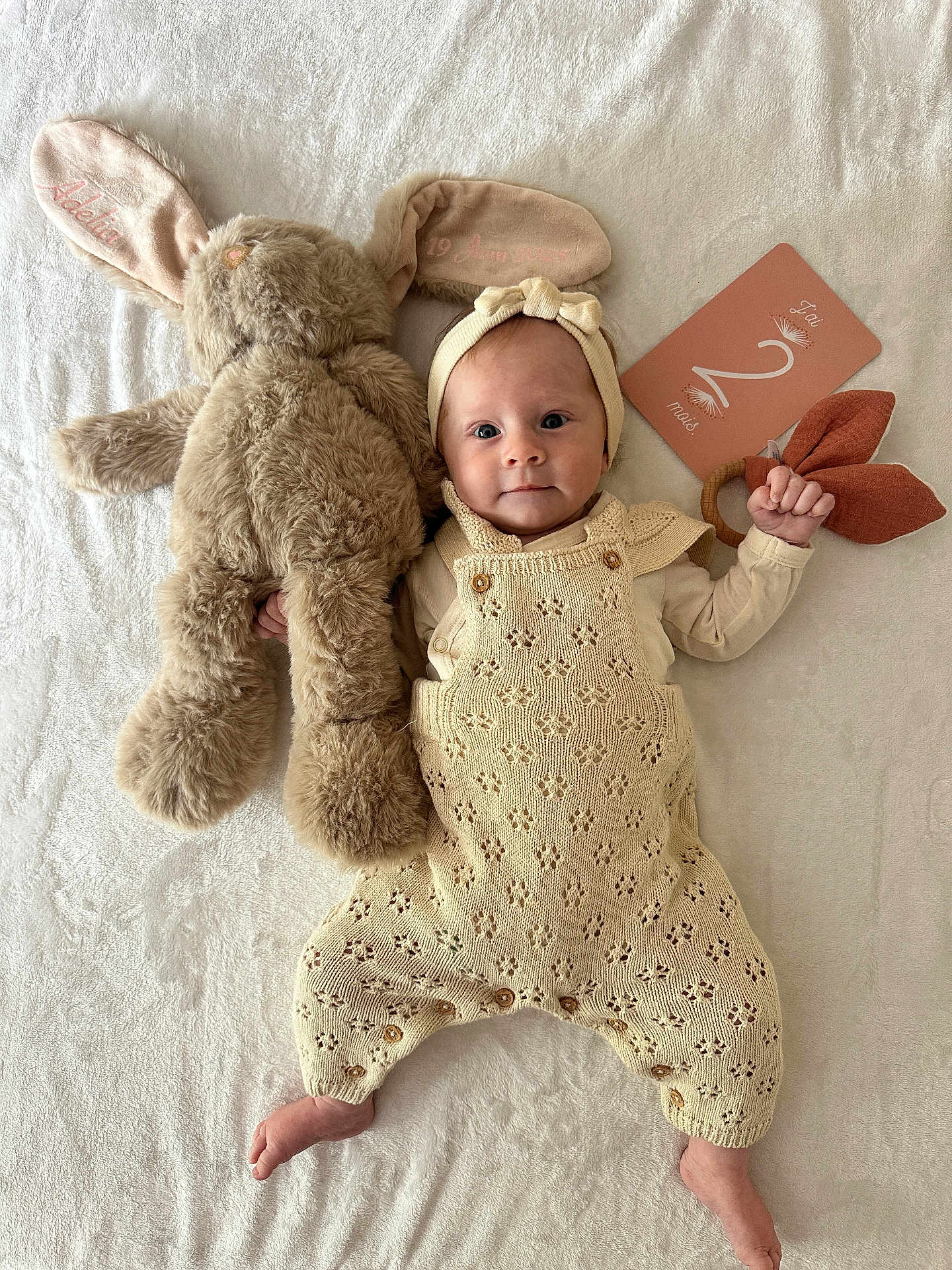 Adelia participe au concours pour gagner de l'argent avec cette photo : baby, infant, knitted_clothing, headband, stuffed_animal, bunny, toy, milestone_card, blanket, soft_texture, lying_down, hand, foot, cute, smile, face, child, indoor, portrait, laying