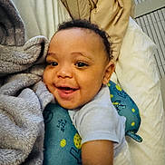 Aspen And Denver is registered to the contest to win money with this photo: baby, smiling, face, blanket, pillow, bed, infant, child, happy, cozy, indoors, soft_texture, cuddling, blue, clothing, person, cute, portrait, young, sleeping_area