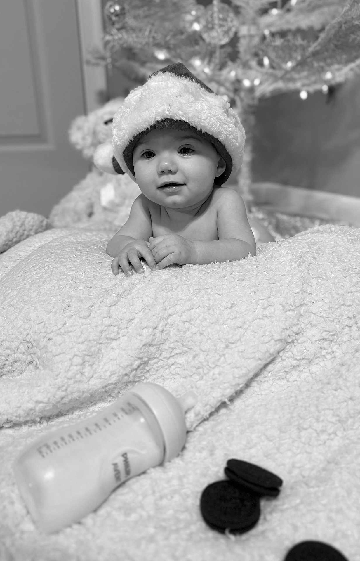 Selena is registered to the contest to win money with this photo: baby, santa_hat, blanket, bottle, cookies, indoors, holiday, christmas_tree, toys, smiling, child, cute, portrait, soft_lighting, black_and_white, cozy, infant, face, hands, floor