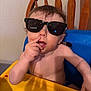 toddler, child, sunglasses, high_chair, yellow_tray, shirtless, indoor, furniture, wooden_chair, curly_hair, messy, cute, baby, hand, face, expression, mouth, skin, person, seat