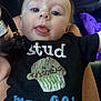 Liam is registered to the contest to win money with this photo: baby, child, face, tongue, black_shirt, text_on_clothing, muffin_graphic, close_up, expression, curious, person, infant, cute, playful, blue_eyes, skin, hair, sitting, clothing, indoor