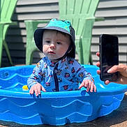 Liam joined the competition — help win amazing prizes! toddler, child, pool, blue_pool, hat, frog_hat, swimwear, outdoor, sunlight, plastic_chair, green_chair, smartphone, hand, water, summer, curious, baby, playful, backyard, concrete