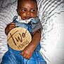Auguste is registered to the contest to win money with this photo: baby, infant, two_months, denim_overalls, clothing, hand, face, eyes, skin, blanket, white, texture, milestone_sign, wooden_sign, expression, curious, cute, portrait, indoor, child