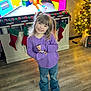 child, girl, pigtails, smiling, purple_shirt, jeans, brown_shoes, living_room, christmas_tree, stockings, flooring, cabinet, television, holiday_decor, ornaments, festive, home_interior, happy, person, cute