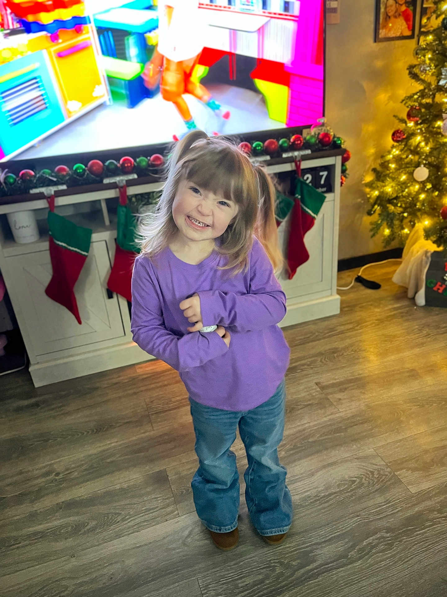 Raylee is registered to the contest to win money with this photo: child, girl, pigtails, smiling, purple_shirt, jeans, brown_shoes, living_room, christmas_tree, stockings, flooring, cabinet, television, holiday_decor, ornaments, festive, home_interior, happy, person, cute