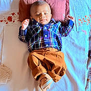 Trishiv is registered to the contest to win money with this photo: baby, infant, plaid_shirt, brown_pants, pillow, bed, blanket, footwear, sleepwear, child, relaxing, indoors, cute, portrait, smiling, person, young, cozy, comfortable, sleepy