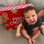 Sophie is registered to the contest to win money with this photo: baby, child, infant, polka_dot, black_clothing, couch, sofa, holiday, christmas, gift_box, gingerbread, smiling, happy, person, indoors, hand, feet, cute, red_bracelet, face
