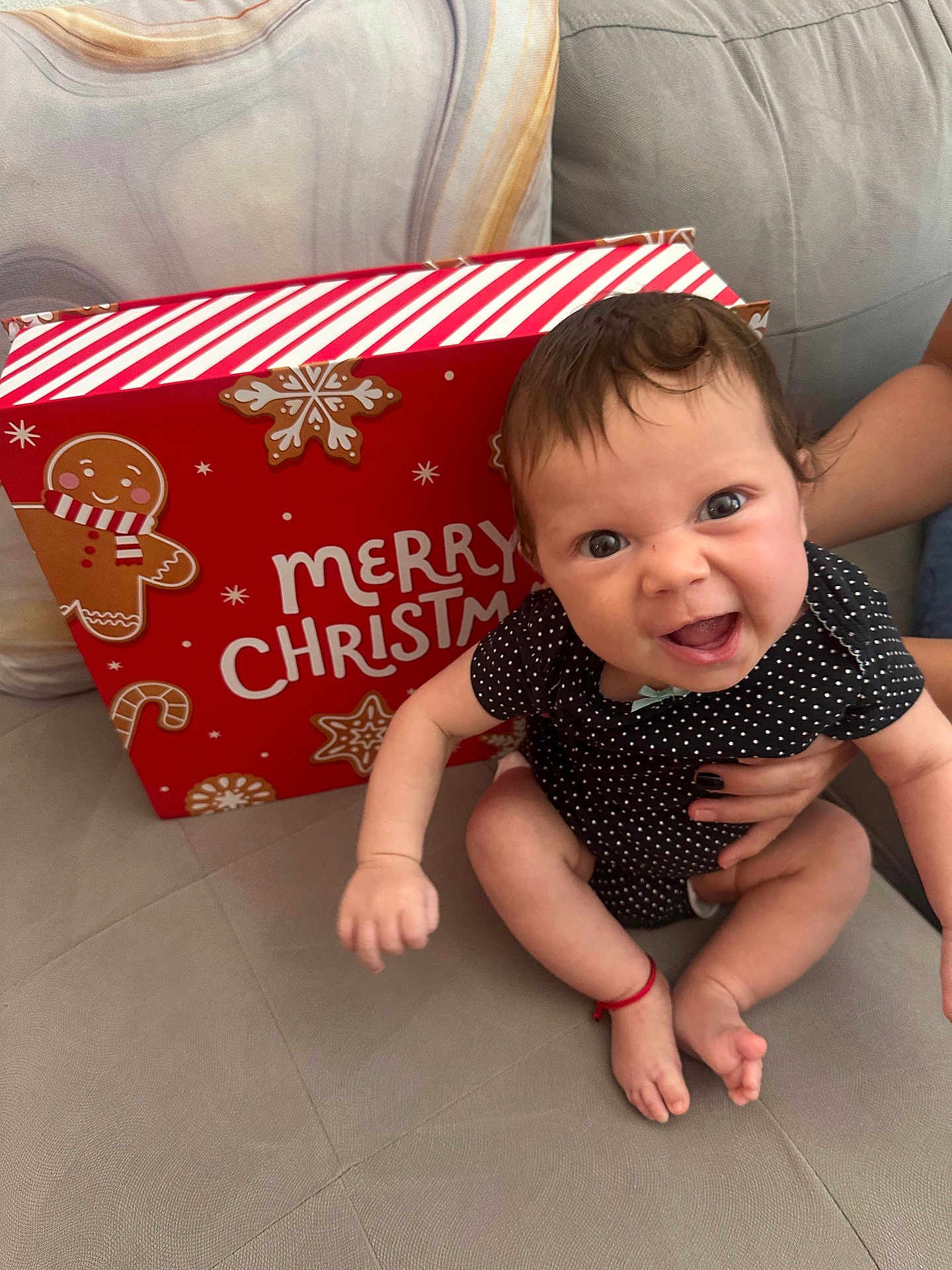 Sophie is registered to the contest to win money with this photo: baby, child, infant, polka_dot, black_clothing, couch, sofa, holiday, christmas, gift_box, gingerbread, smiling, happy, person, indoors, hand, feet, cute, red_bracelet, face