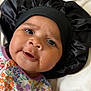Charlotte is registered to the contest to win money with this photo: baby, infant, face, eyes, smile, bonnet, hat, black, floral_outfit, colorful, clothing, portrait, closeup, cute, child, headwear, laying_down, soft, fabric, skin