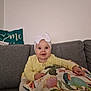 baby, child, headband, bow, yellow_clothing, couch, pillow, floral_pattern, smiling, happy, indoor, person, seated, cute, home, infant, face, wide_eyes, plush, cozy