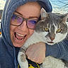 cat, person, selfie, portrait, outdoor, winter, hoodie, glasses, smiling, open_mouth, gray_cat, white_fur, tabby, whiskers, yellow_eyes, hugging, closeup, sky, trees, happy