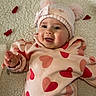 baby, child, smiling, hat, pom_poms, heart_pattern, clothing, blanket, rose_petals, soft_texture, cute, happy, infant, portrait, warm_clothing, indoor, lying_down, face, hand, cozy
