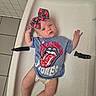 baby, infant, child, bow, tshirt, rolling_stones, changing_table, diaper, tile_floor, tile_wall, expression, cute, clothing, person, indoor, laying, headband, skin, foot, hand