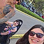 woman, man, child, toddler, selfie, outdoor, bicycle, child_seat, tattoo, sunglasses, cap, smile, family, road, trees, car, parking_lot, helmet, casual_clothing, sunlight