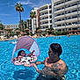 baby, man, pool, floatie, water, sunshade, tattoo, sunglasses, resort, palm_tree, swimming, summer, vacation, sunbathing, people, blue_sky, building, umbrella, relaxation, outdoor