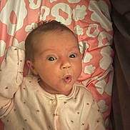 Emily is registered to the contest to win money with this photo: baby, infant, face, expression, onesie, blanket, pattern, pink, white, curly_hair, lying_down, cute, child, small, portrait, soft_lighting, indoors, sleepwear, raised_hand, wide_eyes