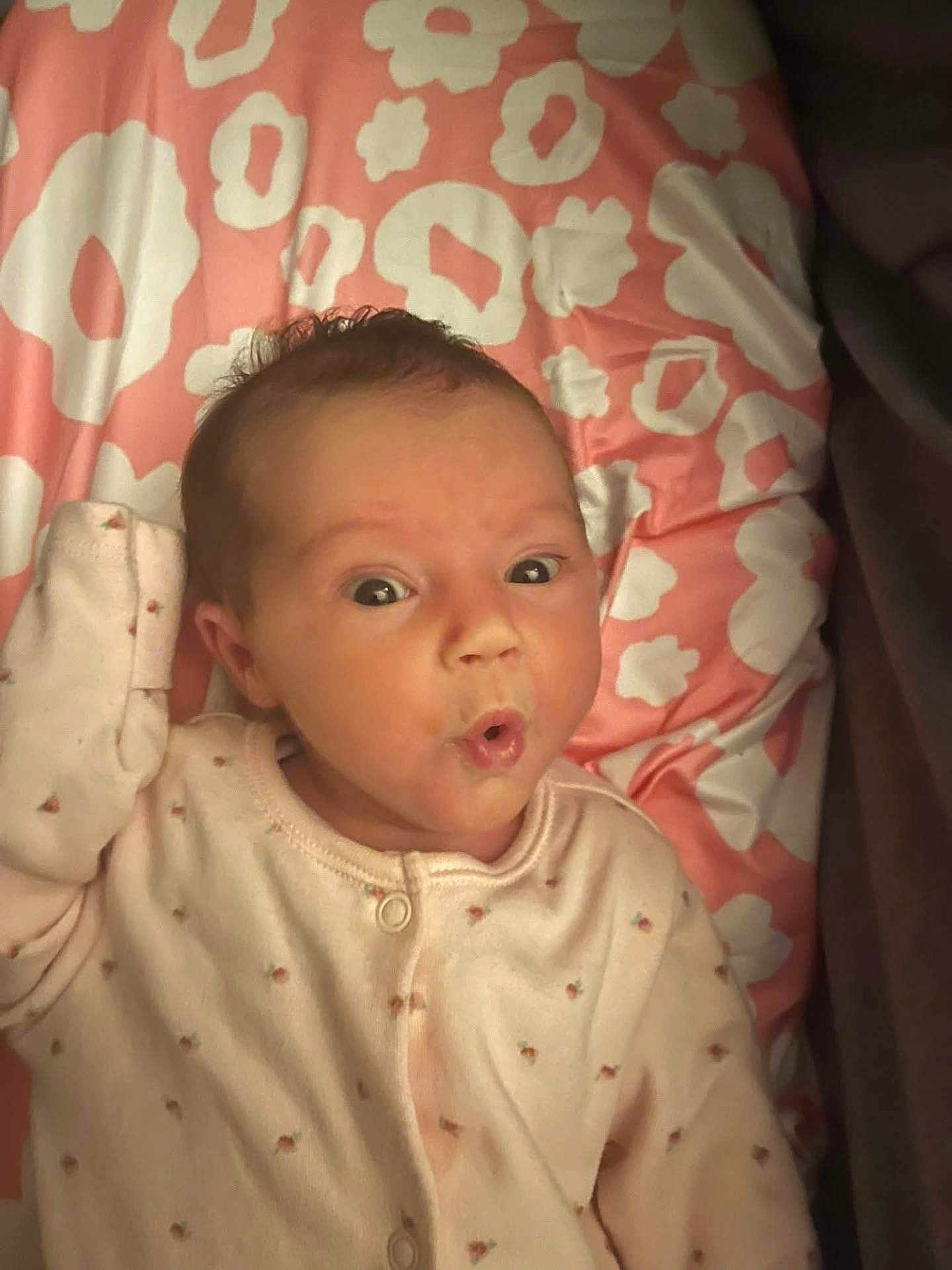 Emily is registered to the contest to win money with this photo: baby, infant, face, expression, onesie, blanket, pattern, pink, white, curly_hair, lying_down, cute, child, small, portrait, soft_lighting, indoors, sleepwear, raised_hand, wide_eyes