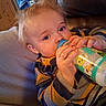 toddler, child, baby_bottle, drinking, striped_clothing, blonde_hair, indoor, cozy, soft_furniture, leather_chair, hand, face, blue_eyes, casual_wear, relaxing, portrait, milk, cute, young_child, home