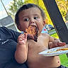 toddler, child, food, meat, plate, outdoor, daylight, person, eating, shirtless, grass, hand, face, black_pants, holding, summer, nature, picnic, happy, casual