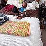 Landon is registered to the contest to win money with this photo: baby, bed, blanket, blue_bib, brown_bedspread, child, clothing, cotton_bedspread, furniture, happy, head, indoors, pillow, quilt, room, sitting, smiling, toy, wooden_bedframe, young_child