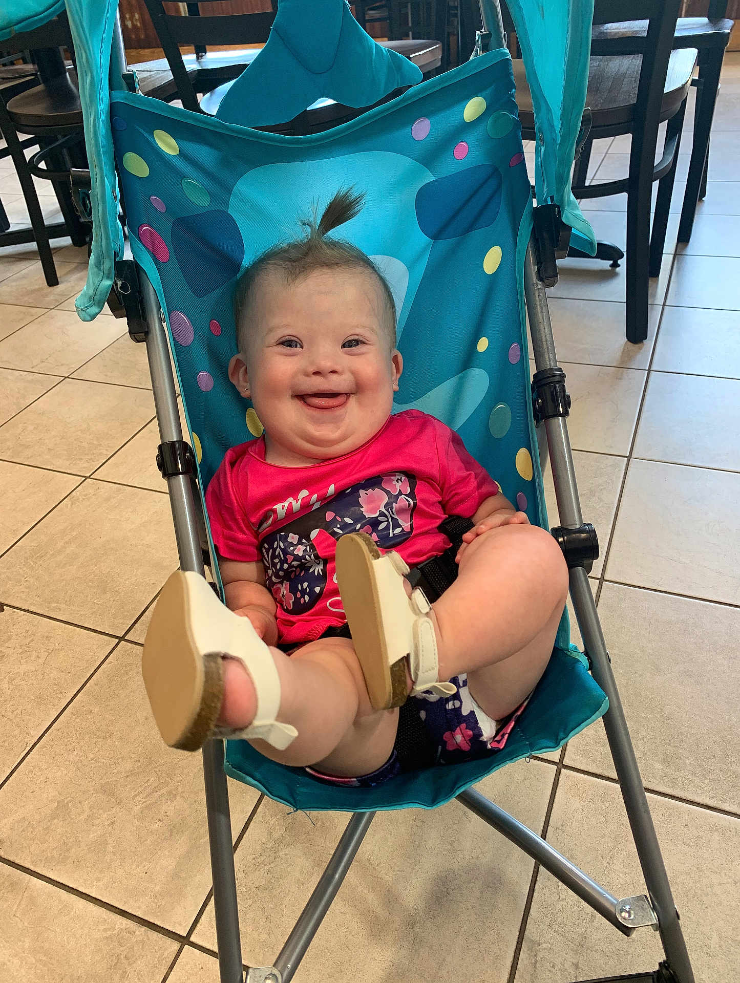 Lakelynn is registered to the contest to win money with this photo: baby, child, smiling, stroller, indoor, floor, tiles, shoes, happy, tongue_out, pink_shirt, blue_stroller, hair_tuft, furniture, chair, seat, playful, cute, person, infant