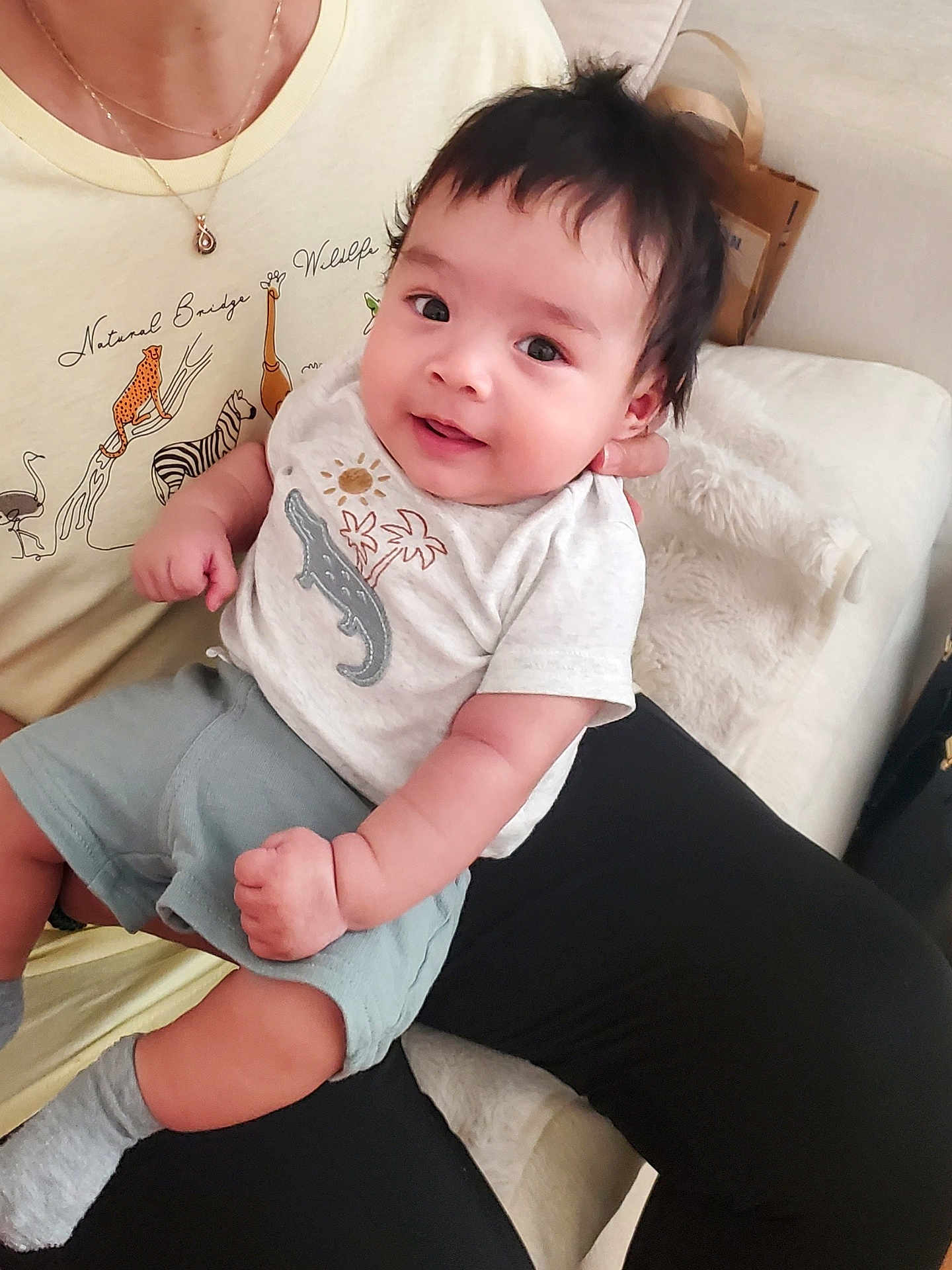 Leonidas joined the competition — help win amazing prizes! baby, child, person, infant, smiling, sitting, clothing, shorts, shirt, hand, face, adult, animal_print, necklace, indoor, couch, blanket, black_hair, skin, cute