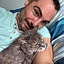 animal, bed, casual_clothing, cat, comfort, cozy, cute, facial_hair, friendship, gray_cat, human, indoor, man, pet, portrait, relaxing, selfie, short_hair, smile, striped_bedding