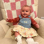 Emery is registered to the contest to win money with this photo: baby, child, smiling, denim_overalls, floral_skirt, socks, armchair, blanket, crocheted, pink, white, beige, indoor, cute, happy, person, portrait, clothing, cozy, infant