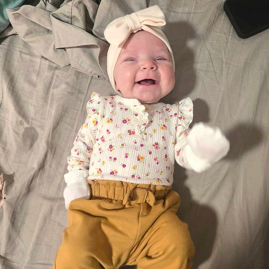 Ensley joined the competition — help win amazing prizes! baby, bed, blanket, bow, child, cute, floral_clothing, happy, headband, indoors, infant, lying_down, mittens, pants, person, phone, portrait, small_hand, smiling, soft_fabric