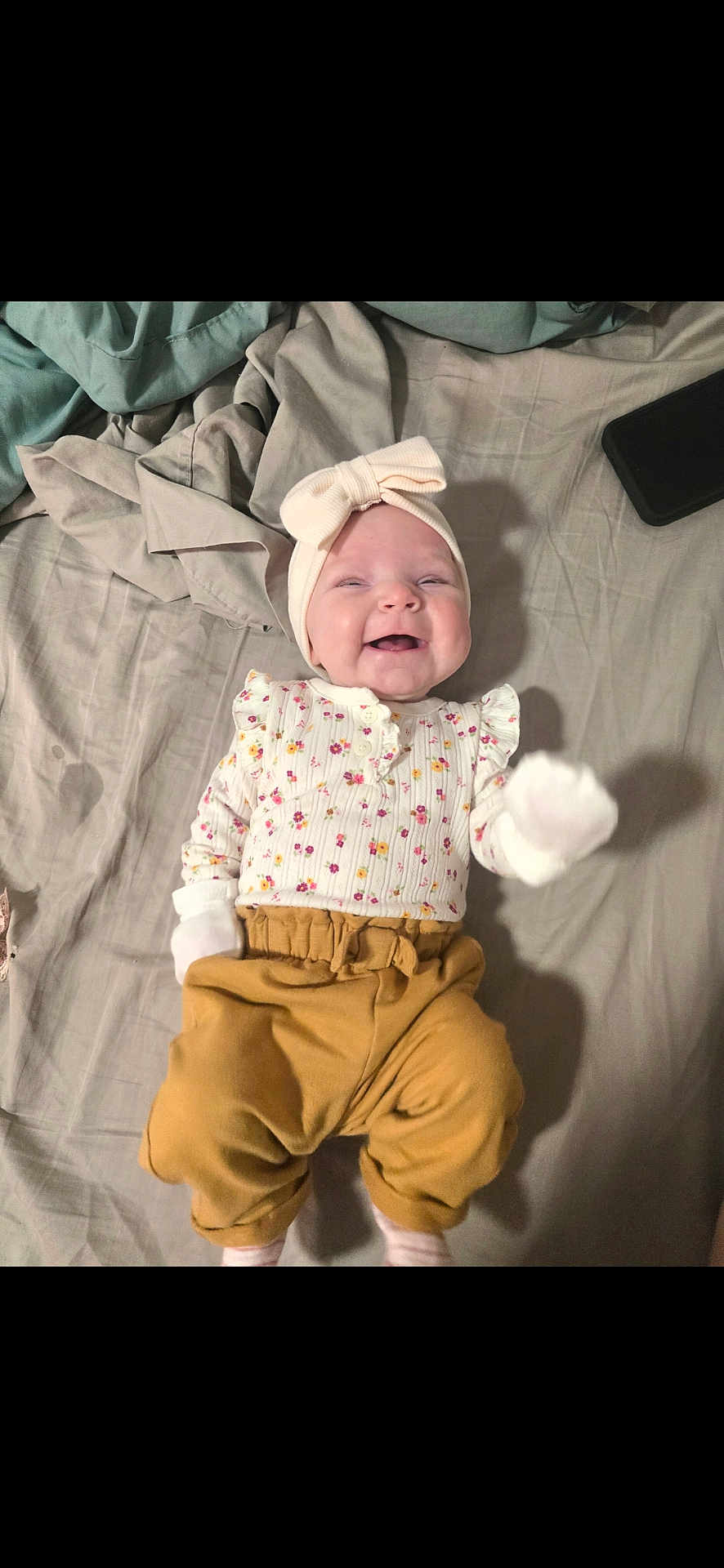 Ensley joined the competition — help win amazing prizes! baby, infant, smiling, headband, bow, floral_clothing, mittens, pants, bed, blanket, phone, happy, cute, lying_down, soft_fabric, child, person, indoors, portrait, small_hand