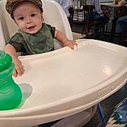Tatem is registered to the contest to win money with this photo: baby, high_chair, bib, cap, smile, child, green_sippy_cup, restaurant, table, chair, adult, person, indoor, casual_clothing, floor, window_blinds, glasses, conversation, furniture, happy