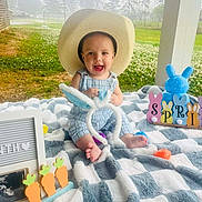 Tatem is registered to the contest to win money with this photo: baby, cowboy_hat, blanket, outdoor, grass, trees, toy, bunny_ears, carrots, easter_decor, smiling, child, cute, plush, colorful, sunlight, person, portrait, happy, nature