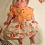 Sienna is registered to the contest to win money with this photo: baby, infant, child, headband, bow, orange_clothing, floral_skirt, lying_down, hands, bed, blanket, cute, skin, foot, hand, person, indoors, soft_lighting, portrait, apparel