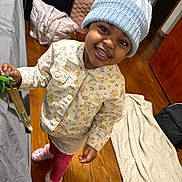 Kimyah is registered to the contest to win money with this photo: child, toddler, smiling, hat, knit_hat, jacket, floral_pattern, pink_pants, indoor, wooden_floor, bed, blanket, toy, pacifier, happy, person, footwear, warm_clothing, home, cute