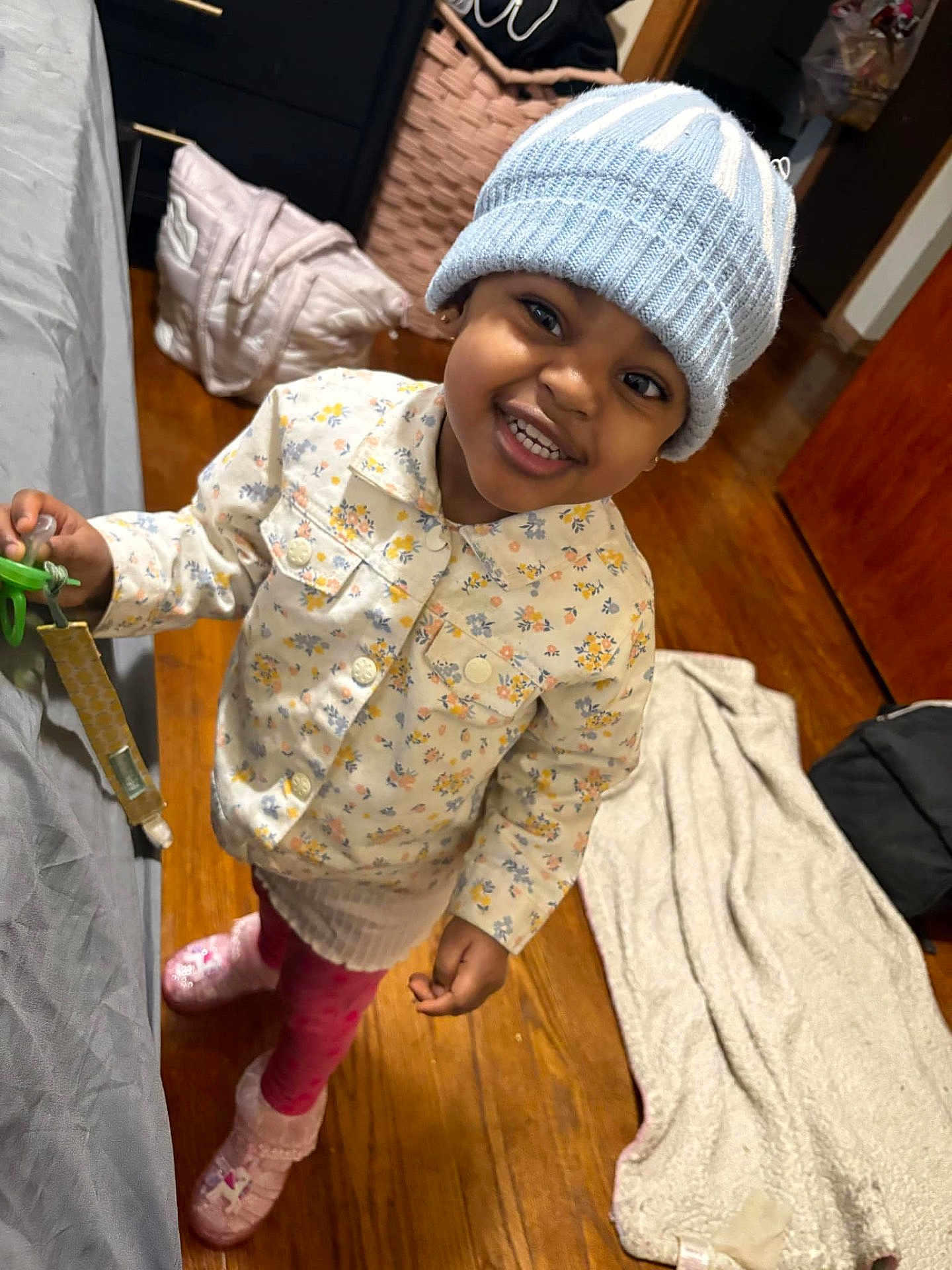 Kimyah is registered to the contest to win money with this photo: child, toddler, smiling, hat, knit_hat, jacket, floral_pattern, pink_pants, indoor, wooden_floor, bed, blanket, toy, pacifier, happy, person, footwear, warm_clothing, home, cute